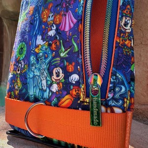 Może przedstawiać: Kolorowa saszetka na zamek błyskawiczny z nadrukiem o tematyce Halloween z postaciami z kresk&oacute;wek. Saszetka ma jasnopomarańczowy pasek i tęczowy zamek błyskawiczny. Na suwaku znajduje się napis "handmade".