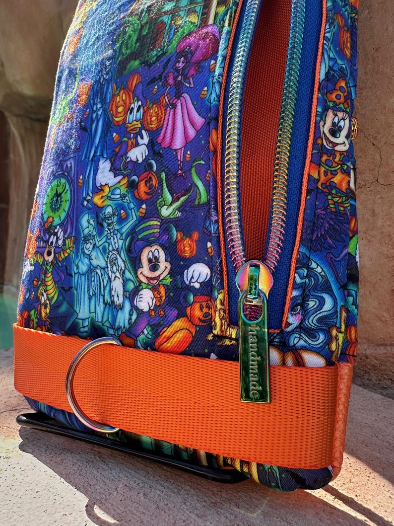 Może przedstawiać: Kolorowa saszetka na zamek błyskawiczny z nadrukiem o tematyce Halloween z postaciami z kresk&oacute;wek. Saszetka ma jasnopomarańczowy pasek i tęczowy zamek błyskawiczny. Na suwaku znajduje się napis "handmade".