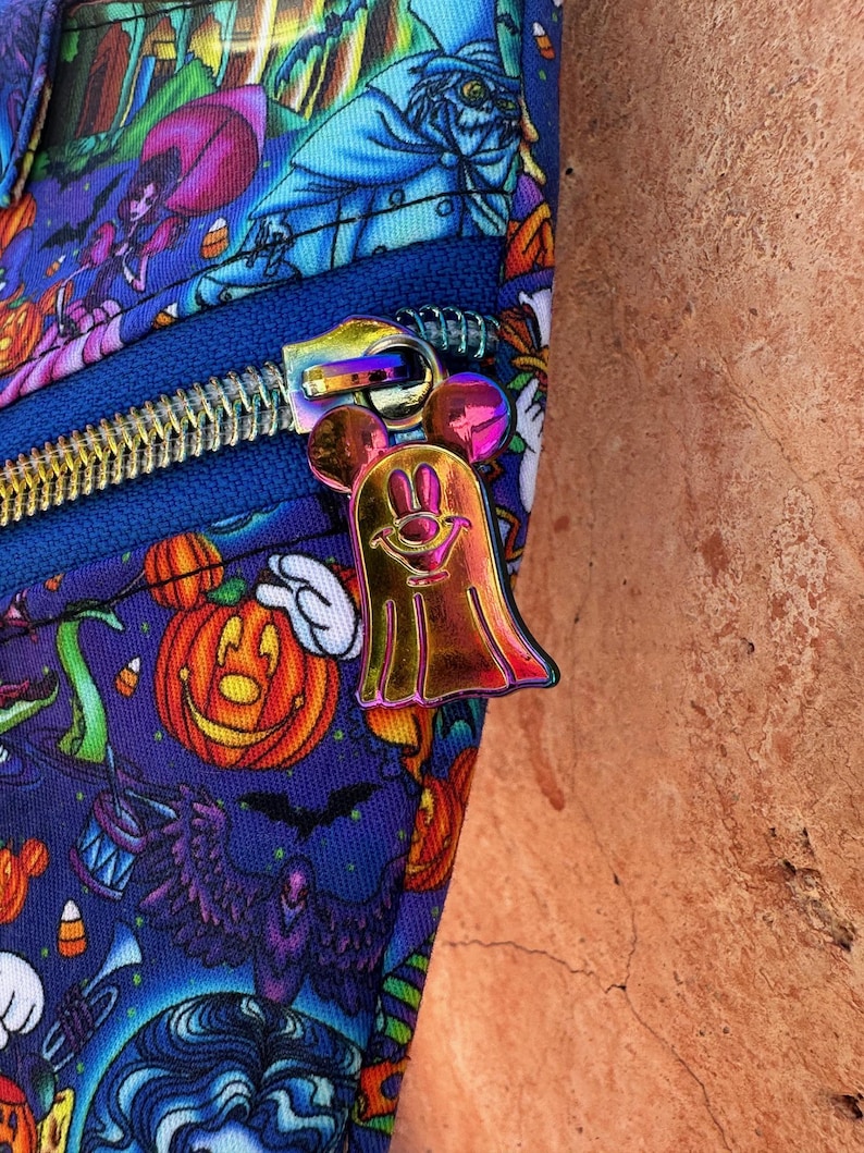 Może przedstawiać: Zbliżenie niebieskiej tkaniny z nadrukiem o tematyce Halloween, przedstawiającym dynie, duchy i inne upiorne postacie. Widoczny jest niebieski zamek błyskawiczny z metalicznym, iryzującym uchwytem zamka błyskawicznego w kształcie ducha Myszki Miki.