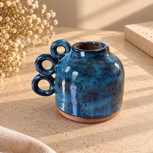 May include: A small, round ceramic vase with a deep blue, mottled glaze and a smooth, dark brown base. The vessel features a unique handle design consisting of three vertically stacked, circular rings attached to one side of the body.