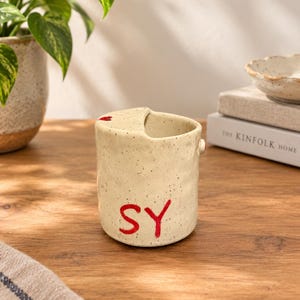 May include: A cylindrical ceramic mug with a speckled cream finish and an irregular, wavy rim. The exterior features the red painted letters SY on the front and a small red heart detail on the top edge. The mug has a smooth, glossy glaze.