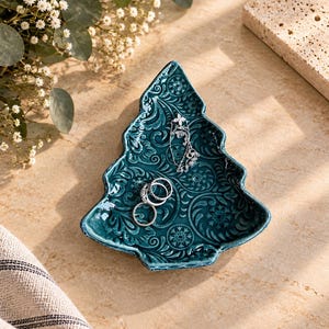 May include: A teal ceramic trinket dish shaped like a Christmas tree, featuring an embossed pattern of swirling leaves and snowflakes. The dish holds three silver-toned rings and a delicate silver chain necklace with a small floral pendant.