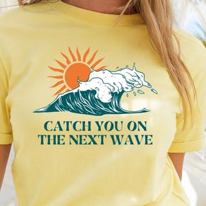 May include: Yellow t-shirt with a graphic of a sun, ocean wave, and the text "CATCH YOU ON THE NEXT WAVE". The shirt has short sleeves and a crew neck. The design features orange, white, and teal colors.