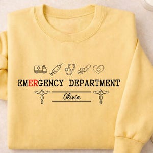 May include: A pale yellow crew neck sweatshirt featuring the text "EMERGENCY DEPARTMENT" in black, with medical symbols above. The name "Olivia" is printed below. The sweatshirt is a light yellow colour.