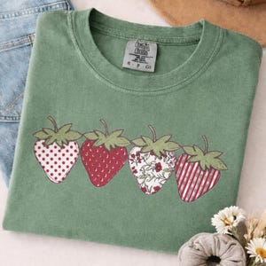 Puede incluir: Una camiseta verde salvia con un diseño de fresas bordadas. Las fresas tienen diferentes patrones, incluyendo lunares, rojo liso, floral y rayas. La camiseta está doblada junto a unos vaqueros azules y un sombrero de paja.