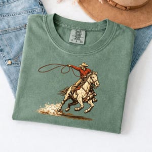 May include: Sage green t-shirt featuring a cowboy on a horse, roping. The cowboy wears a red shirt and a hat. The horse is tan and brown. The shirt is folded on top of blue jeans.
