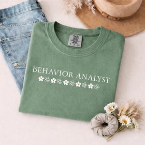 Peut inclure: Un t-shirt à col rond vert sauge avec le texte blanc BEHAVIOR ANALYST centré au-dessus d'une rangée horizontale de petites illustrations de fleurs blanches et orange. Le t-shirt porte une étiquette de marque Comfort Colors à l'intérieur.