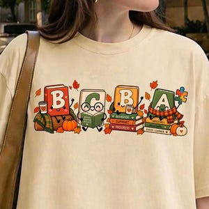 May include: A cream-coloured short-sleeve t-shirt featuring a graphic print of four anthropomorphic books labelled B, C, B, and A. The books are decorated with autumn leaves, pumpkins, and a puzzle piece, with small coffee cups and text accents.