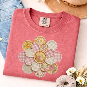 Könnte beinhalten: Ein korallenfarbenes T-Shirt mit einer großen, zentralen Blumengrafik, die aus verschiedenen stoffartigen Flicken mit Blumen-, Vichy- und Unimustern besteht. Das T-Shirt hat einen Rundhalsausschnitt und ein Comfort Colors Markenetikett am Innenkragen.