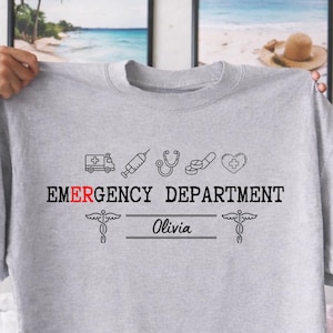May include: A heather gray t-shirt with the words "EMERGENCY DEPARTMENT" in black, with a red accent. The shirt features medical-themed icons and the name "Olivia" below. The design includes a caduceus symbol on each side.