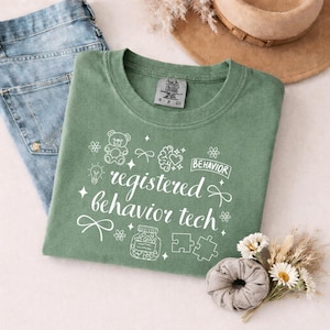 May include: A sage green t-shirt featuring white line art graphics including a teddy bear, a brain with a heart, a lightbulb, a jar of pills, puzzle pieces, and bows. The text reads registered behavior tech and behavior.