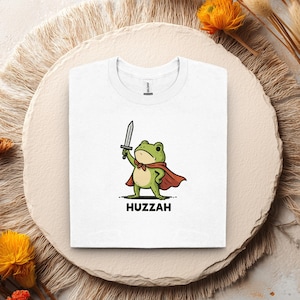 May include: White t-shirt featuring a cartoon frog character wearing a red cape and holding a sword. The frog has a green body, a red bow tie, and the word "Huzzah" is printed below the frog in black letters.