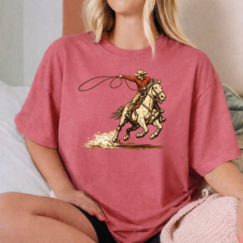 May include: A dusty rose short-sleeved t-shirt featuring a graphic illustration of a cowboy in a red shirt and hat riding a white horse while swinging a lasso. The horse is depicted in motion, kicking up dust from the ground below its hooves.