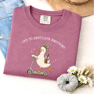 May include: A mauve crewneck t-shirt featuring a graphic of a white duck wearing a green headband, riding a light green scooter, and holding an iced coffee. The text above the duck reads OFF TO REGULATE EMOTIONS in white capital letters.