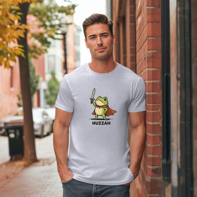 May include: Light gray t-shirt featuring a cartoon frog warrior holding a sword and wearing a red cape. The word "Huzzah" is printed below the frog. The shirt is a crew neck style.