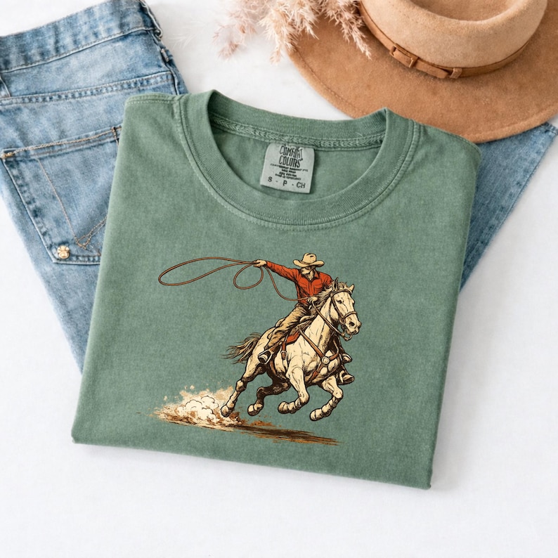 May include: Sage green t-shirt featuring a cowboy on a horse, roping. The cowboy wears a red shirt and a hat. The horse is tan and brown. The shirt is folded on top of blue jeans.