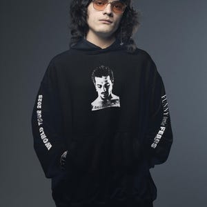 K&ouml;nnte beinhalten: Schwarzer Kapuzenpullover mit Portr&auml;t und Text. Auf der Vorderseite des Kapuzenpullovers ist ein Schwarz-Wei&szlig;-Bild eines Mannes zu sehen. Die &Auml;rmel tragen den Text "WORLD TOUR 2025" und "FUNNY little FEARS" in Wei&szlig;. Die Person auf dem Bild tr&auml;gt orange get&ouml;nte Gl&auml;ser.