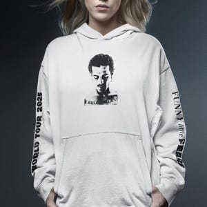 K&ouml;nnte beinhalten: Cremefarbener Kapuzenpullover mit einem Schwarz-Wei&szlig;-Portr&auml;t auf der Vorderseite. Die &Auml;rmel zeigen den Text "WORLD TOUR 2025" und "FUNNY little FEARS" in Schwarz. Der Kapuzenpullover hat eine Vordertasche und eine Kapuze mit Kordelzug.