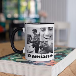 以下が含まれることがあります： 黒い内側とハンドルが付いた白いセラミックマグカップ。マグカップには、人物の4つの白黒写真と、下に「Damiano」という名前が印刷されています。マグカップは、カラフルな花柄のデザインの上の本の上に置かれています。