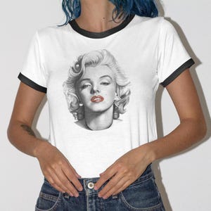 以下が含まれることがあります： 白のTシャツ、黒のトリム付き。マリリン・モンローの白黒ポートレートが特徴です。ポートレートは、赤い口紅と巻き毛のブロンドの髪を持つモンローを示しています。シャツは半袖で、クルーネックです。