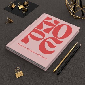May include: A pink journal with the word "POEM" in large coral letters. The text "A place for thoughts and prayers" is below. Gold and black pencils and gold binder clips are nearby.