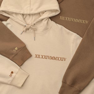May include: Two hooded sweatshirts, one beige and one brown, with embroidered roman numerals and hearts. The beige sweatshirt has the roman numerals "XII.II.IV.MMXXVI" and the brown sweatshirt has the roman numerals "XII.II.IV.MMXXVI".
