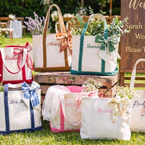 May include: A collection of six canvas tote bags in various colors, each featuring a personalized name embroidered on the front, two sturdy handles, and a decorative patterned ribbon tied to one side, filled with assorted fresh flowers and greenery.
