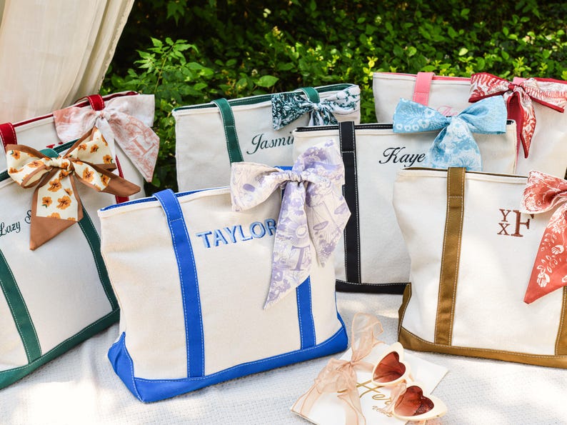 May include: A collection of personalized canvas tote bags in various colors, each adorned with a decorative scarf bow. The bags feature embroidered names or initials, with accents like blue, green, and brown trim. The bags are on a white surface.
