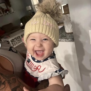 May include: A baby wearing a cream colored knit beanie with a faux fur pom pom and the text "Go Dawgs" embroidered on the front. The baby is wearing a white and red patterned shirt and is smiling.