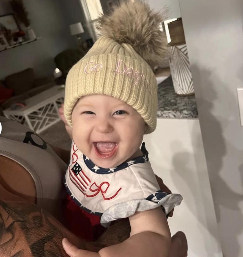 May include: A baby wearing a cream colored knit beanie with a faux fur pom pom and the text "Go Dawgs" embroidered on the front. The baby is wearing a white and red patterned shirt and is smiling.
