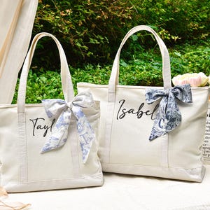 Peut inclure: Deux sacs fourre-tout en toile écrue avec les noms "Tayl" et "Isabel" écrits en script noir. Chaque sac est orné d'un nœud décoratif à motif floral bleu et blanc. Les sacs ont des anses solides et sont posés devant un fond de verdure.