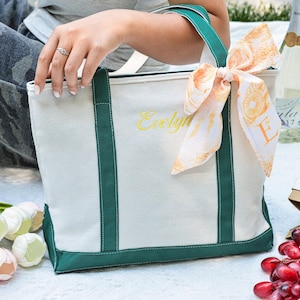 May include: A white canvas tote bag with green trim and handles, embroidered with the name "Evelyn" in yellow. A decorative scarf with an orange pattern is tied to the handle. The bag is on a picnic blanket with flowers, grapes, and a bottle of wine.