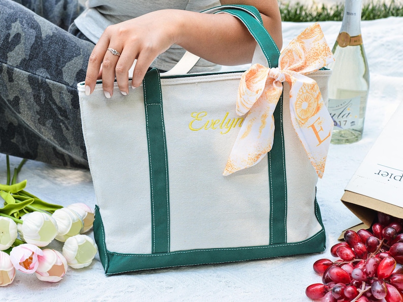 May include: A white canvas tote bag with green trim and handles, embroidered with the name "Evelyn" in yellow. A decorative scarf with an orange pattern is tied to the handle. The bag is on a picnic blanket with flowers, grapes, and a bottle of wine.