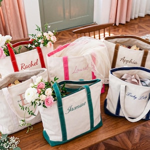 May include: A collection of five off-white canvas tote bags with colored trim and handles. Each bag features custom embroidery in script font with the names Rachel, Laurel, Apphia, Jasmine, and the phrase Lucky Lady in various thread colors.