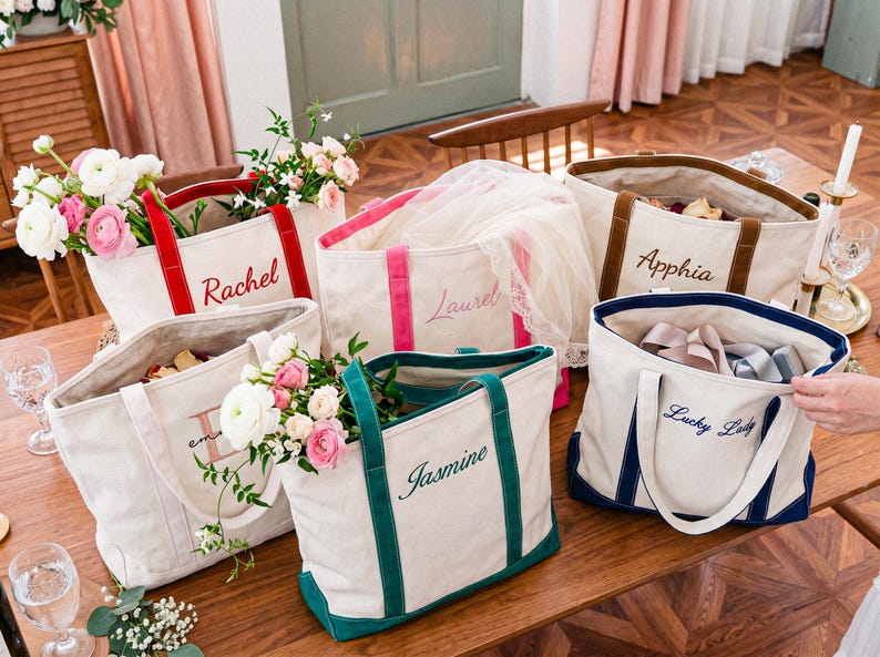 May include: A collection of five off-white canvas tote bags with colored trim and handles. Each bag features custom embroidery in script font with the names Rachel, Laurel, Apphia, Jasmine, and the phrase Lucky Lady in various thread colors.