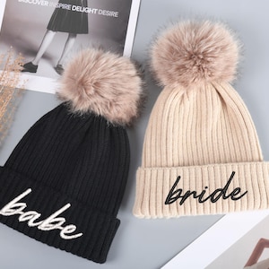 Puede incluir: Dos gorros de punto con pompones de piel sintética. El gorro negro dice "babe" en letras blancas. El gorro beige dice "bride" en letras negras.