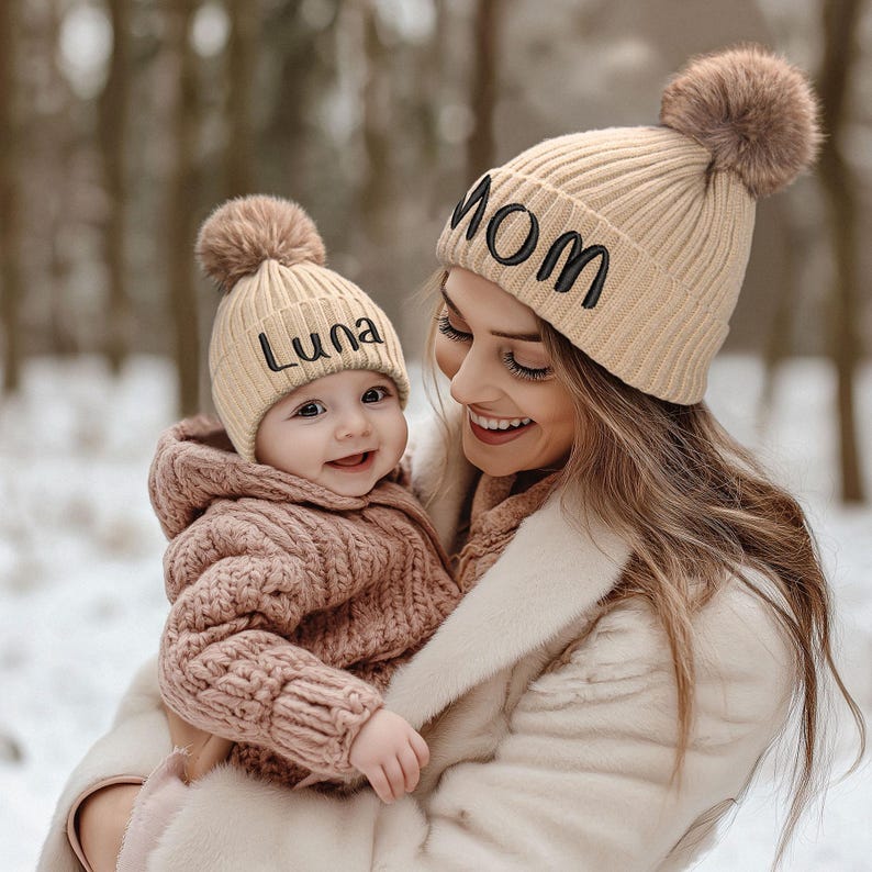 May include: A woman and a baby wearing matching beige knit hats with pom-poms. The woman's hat says "MOM" and the baby's hat says "Luna".
