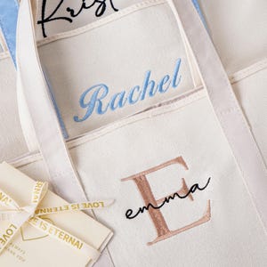 May include: Off-white canvas tote bags featuring personalized embroidery. One bag displays the name Rachel in blue script, another shows a large rose gold letter E with the name emma in black script, and a small card tied with ribbon reads LOVE IS ETERNAL.
