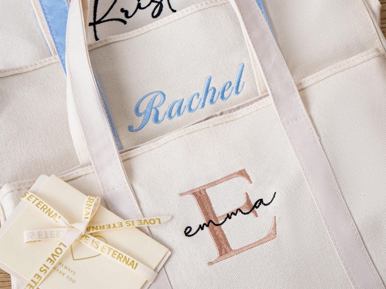 May include: Off-white canvas tote bags featuring personalized embroidery. One bag displays the name Rachel in blue script, another shows a large rose gold letter E with the name emma in black script, and a small card tied with ribbon reads LOVE IS ETERNAL.