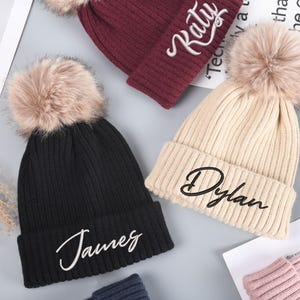 Peut inclure: Trois bonnets en tricot avec des pompons en fausse fourrure. Les bonnets sont noirs, beiges et bordeaux. Chaque bonnet a un nom brodé sur le devant en lettres cursives blanches. Les noms sont "James", "Dylan" et "Katy".