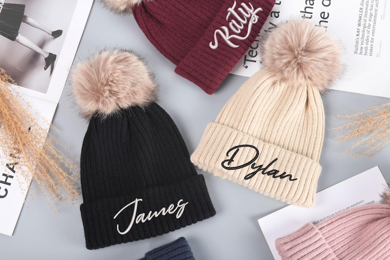 May include: Three knit beanies with faux fur pom poms. The beanies are black, beige, and burgundy. Each beanie has a name embroidered on the front in white cursive lettering. The names are "James", "Dylan", and "Katy".