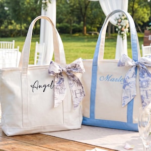 May include: Two canvas tote bags with top handles. The left bag is cream with black text reading "Angel" and a patterned ribbon. The right bag has light blue trim, blue text reading "Marley", and a blue and white patterned ribbon tied to the handle.
