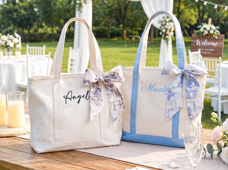 May include: Two canvas tote bags with top handles. The left bag is cream with black text reading "Angel" and a patterned ribbon. The right bag has light blue trim, blue text reading "Marley", and a blue and white patterned ribbon tied to the handle.