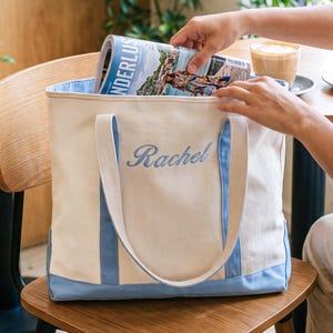 May include: A cream-colored canvas tote bag featuring light blue trim along the bottom and handles. The name Rachel is embroidered in light blue script on the front center of the bag. The bag is open at the top, revealing a light blue interior lining.