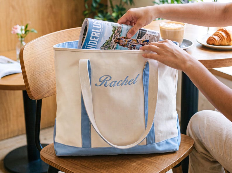 May include: A cream-colored canvas tote bag featuring light blue trim along the bottom and handles. The name Rachel is embroidered in light blue script on the front center of the bag. The bag is open at the top, revealing a light blue interior lining.