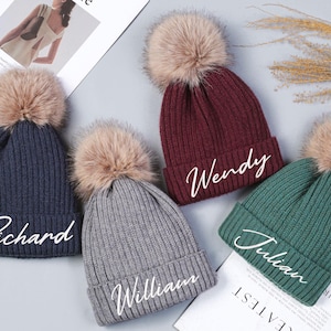 May include: Four knitted beanies with faux fur pom poms and personalized names. The beanies are in navy blue, burgundy, gray, and green.