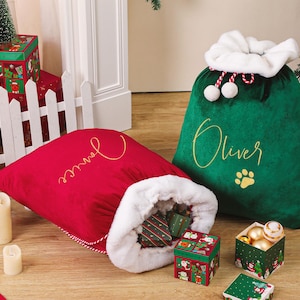 May include: Two plush Christmas gift sacks, one red and one green, sit on a wooden floor. The red sack has the name "Janice" embroidered in gold, and the green sack has "Oliver" embroidered in gold. The sacks are surrounded by wrapped gifts and decorative candles.
