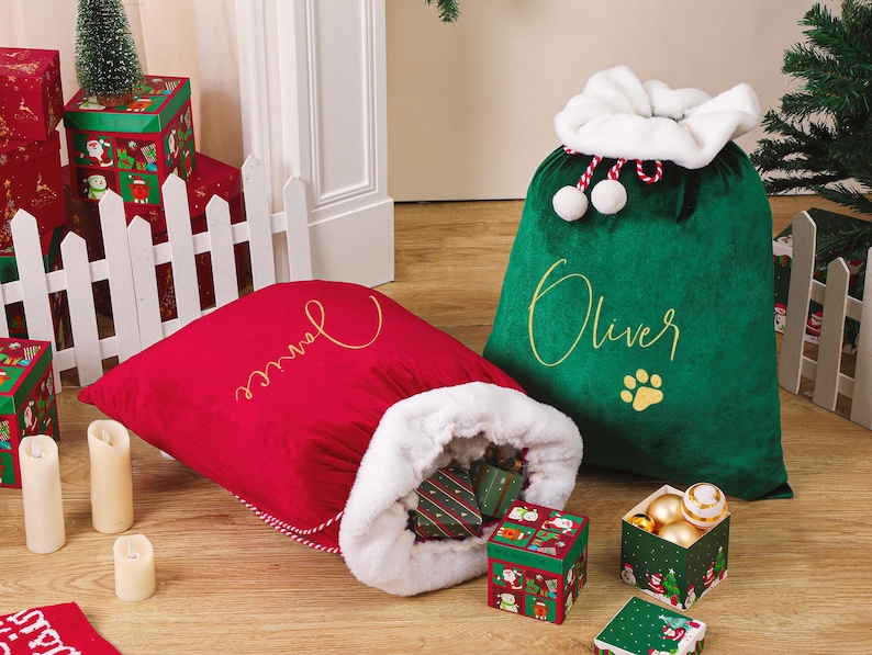 May include: Two plush Christmas gift sacks, one red and one green, sit on a wooden floor. The red sack has the name "Janice" embroidered in gold, and the green sack has "Oliver" embroidered in gold. The sacks are surrounded by wrapped gifts and decorative candles.