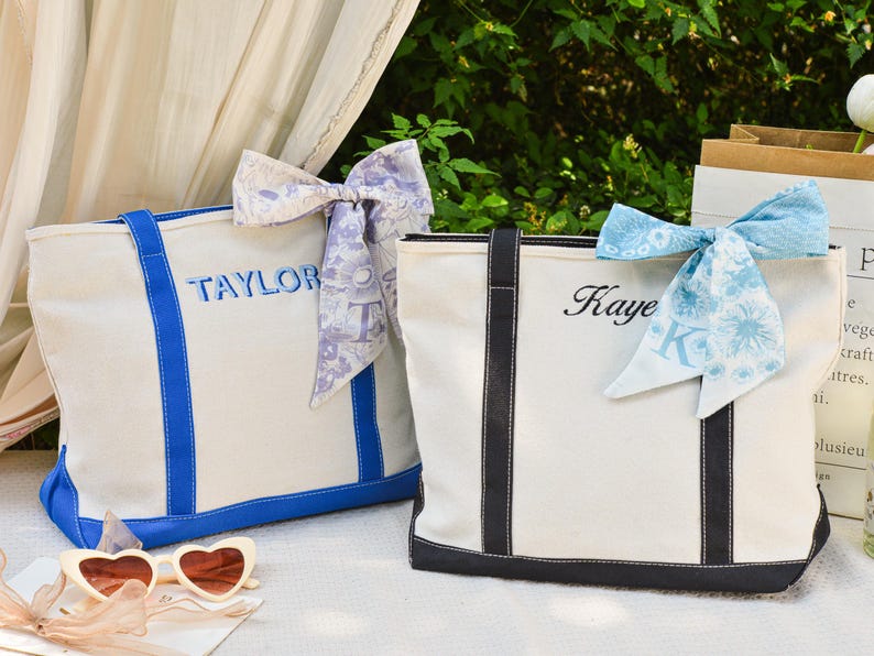 May include: Two canvas tote bags with personalized names embroidered in blue and black. One bag has blue accents and the name "TAYLOR", the other has black accents and the name "Kaye". Each bag is adorned with a decorative scarf.