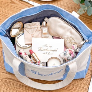 May include: A light blue and white canvas tote bag with the name Rachel embroidered on the front. The bag contains a gold compact mirror, a necklace in a pouch, a perfume bottle, a sleep mask, a floral lipstick, a candle, and a card reading For my Bridesmaid with Love.